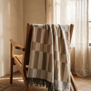 A woven throw blanket with a checkered pattern in beige, brown, and muted blue-gray tones, draped over a wooden chair in a sunlit room with warm neutral walls and curtains.
