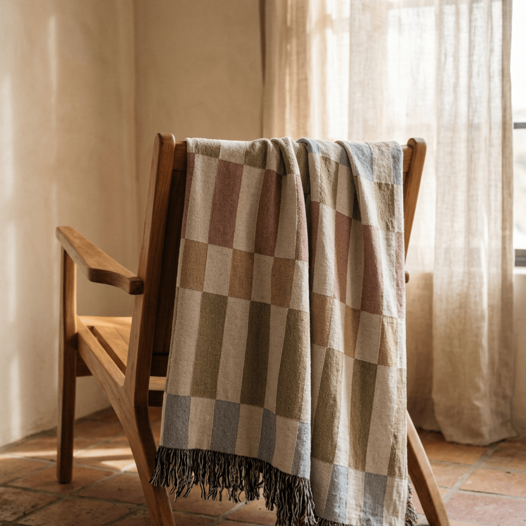 A woven throw blanket with a checkered pattern in beige, brown, and muted blue-gray tones, draped over a wooden chair in a sunlit room with warm neutral walls and curtains.
