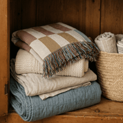 A stack of folded home textiles, including a plaid throw blanket with fringe, a chunky knit cream blanket, a light linen sheet, and a dusty blue quilted blanket, arranged inside a rustic wooden cabinet next to a woven basket.