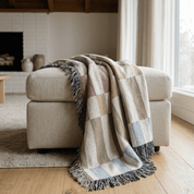 A woven throw blanket with a large check pattern in beige, dusty pink, light blue, and cream, draped over a light beige upholstered ottoman in a bright, modern living room.