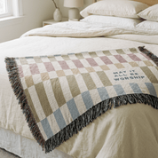 A woven throw blanket with a muted checkered pattern and fringe, draped over a bed. The blanket features the text 'MAY IT ALL BE WORSHIP' in navy blue.