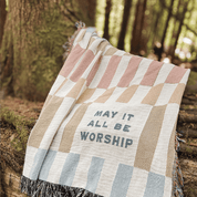 A woven throw blanket with a patchwork pattern in beige, dusty pink, and light blue, featuring the text 'MAY IT ALL BE WORSHIP' and fringed edges, photographed outdoors against a blurred forest background.