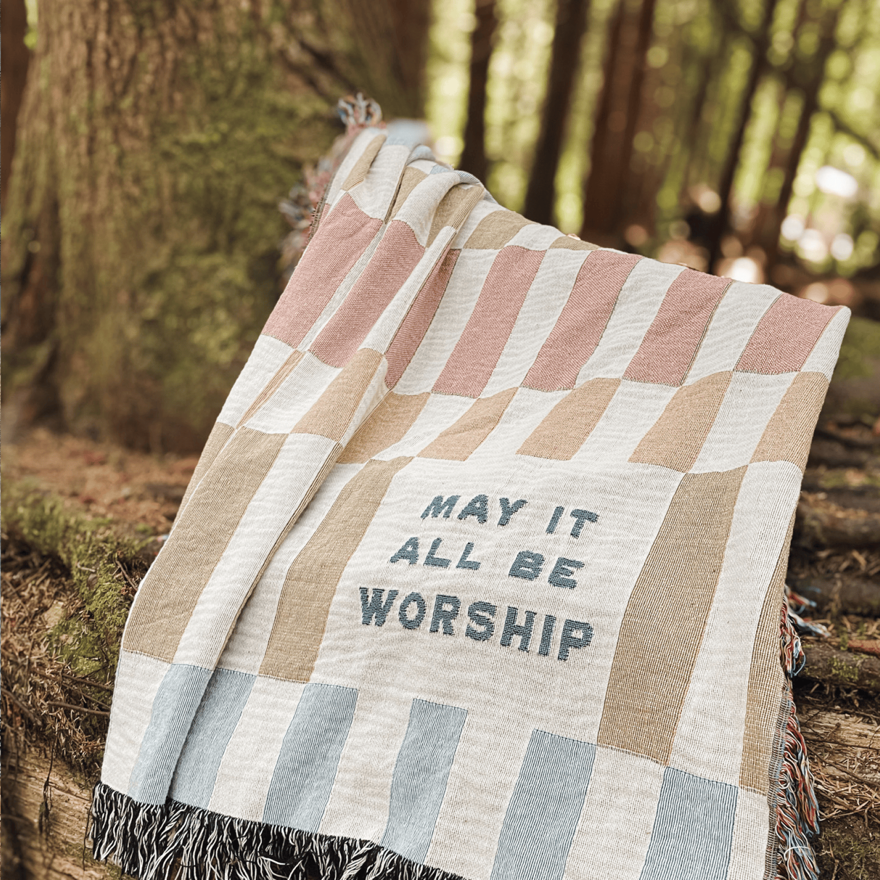 A woven throw blanket with a patchwork pattern in beige, dusty pink, and light blue, featuring the text 'MAY IT ALL BE WORSHIP' and fringed edges, photographed outdoors against a blurred forest background.