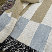 A close-up of a woven patchwork blanket with large blocks of cream, light blue, and tan, finished with black and white fringe.