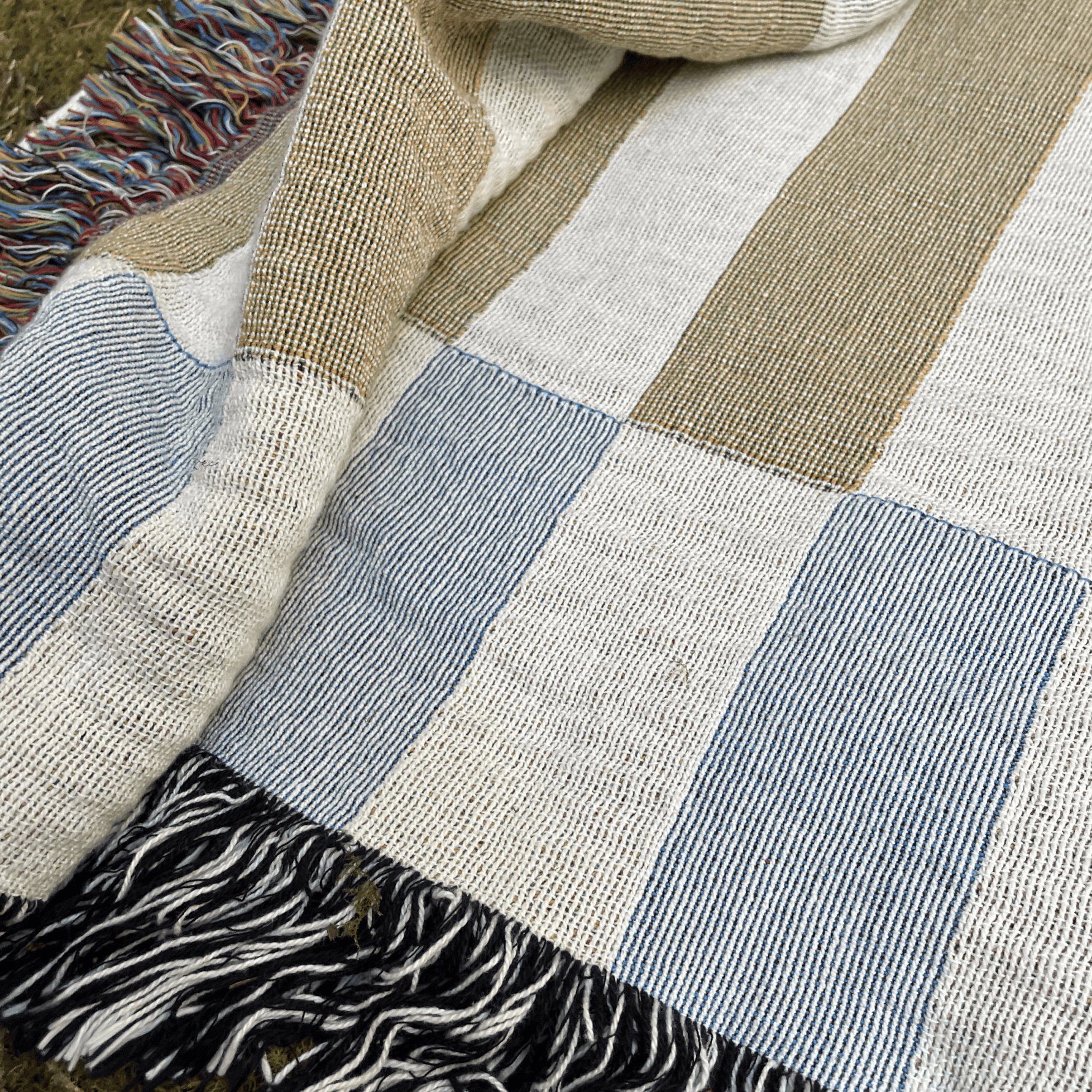 A close-up of a woven patchwork blanket with large blocks of cream, light blue, and tan, finished with black and white fringe.