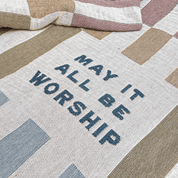 A close-up of a woven textile with the phrase “MAY IT ALL BE WORSHIP” in dark blue lettering, laid over a patchwork design in beige, cream, and muted mauve tones.