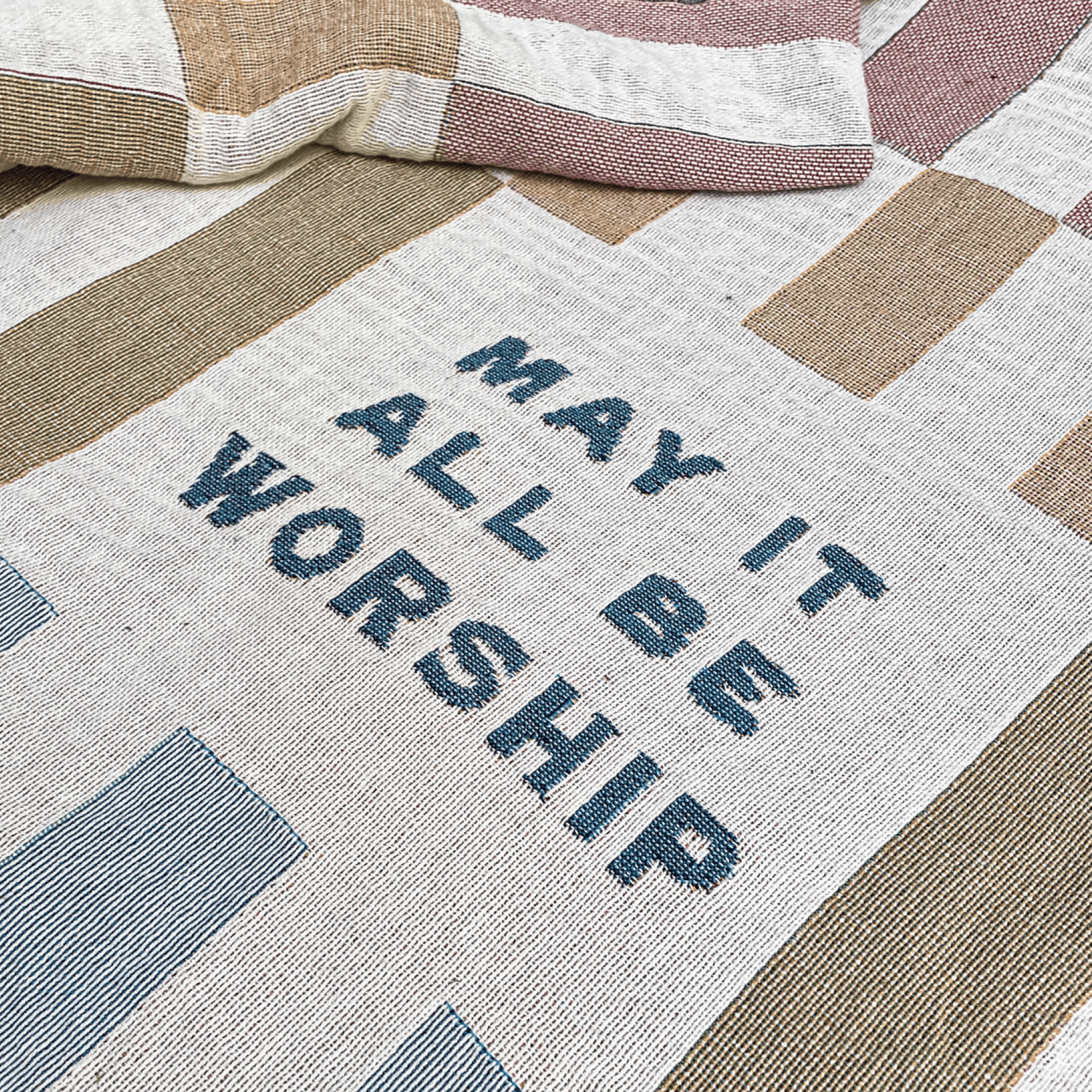 A close-up of a woven textile with the phrase “MAY IT ALL BE WORSHIP” in dark blue lettering, laid over a patchwork design in beige, cream, and muted mauve tones.