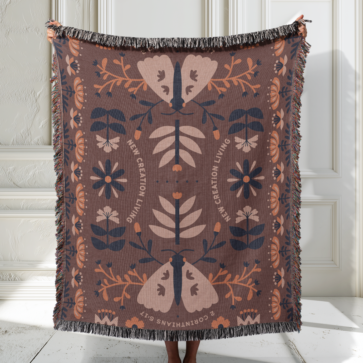 Decorative blanket with floral and butterfly pattern held by a person against a white wall.