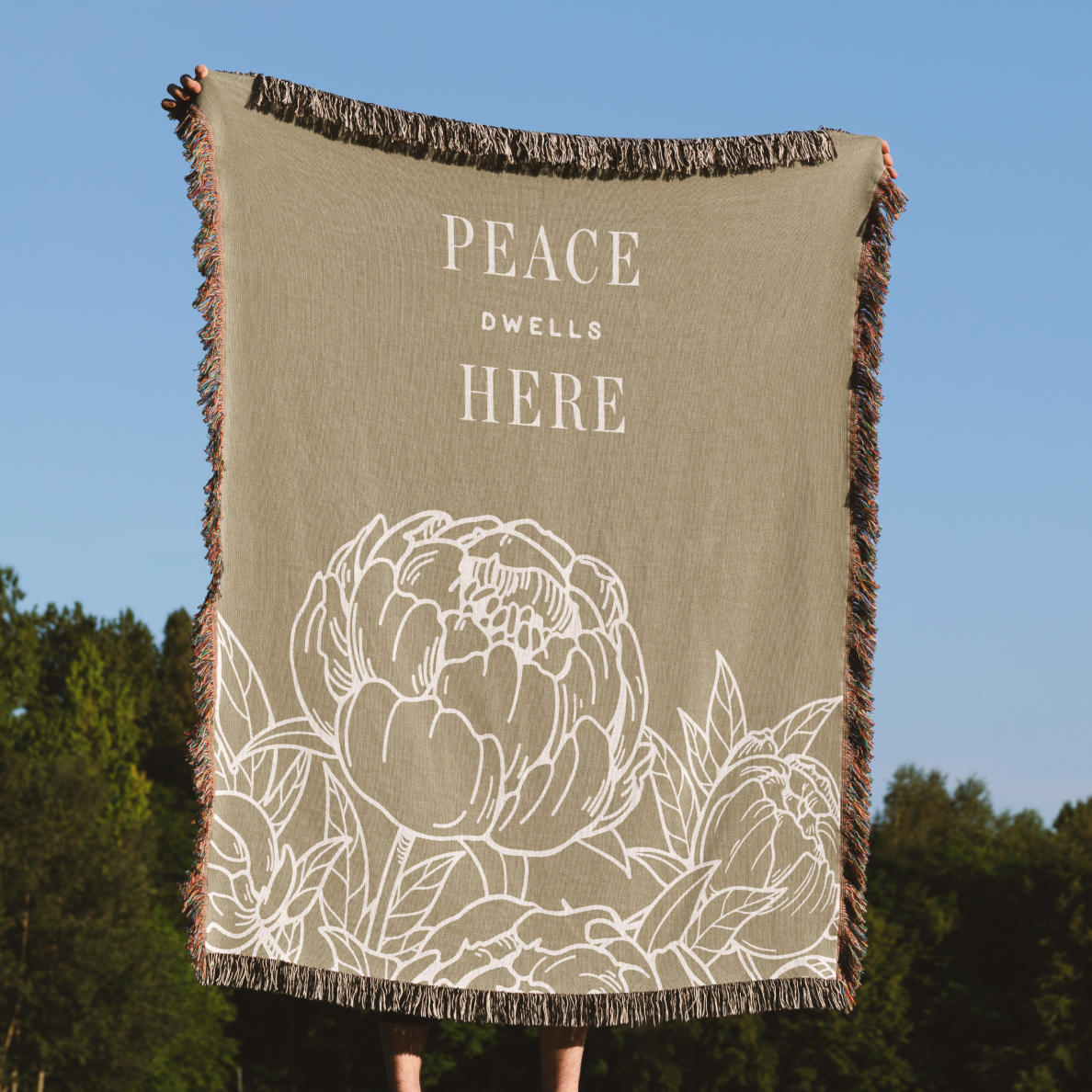 Blanket with 'Peace Dwell Here' text and floral design against a blue sky and trees background
