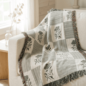 A cream and olive green throw blanket with a floral and striped patchwork pattern and fringed edges, draped over a light beige armchair in a bright, modern living room setting.