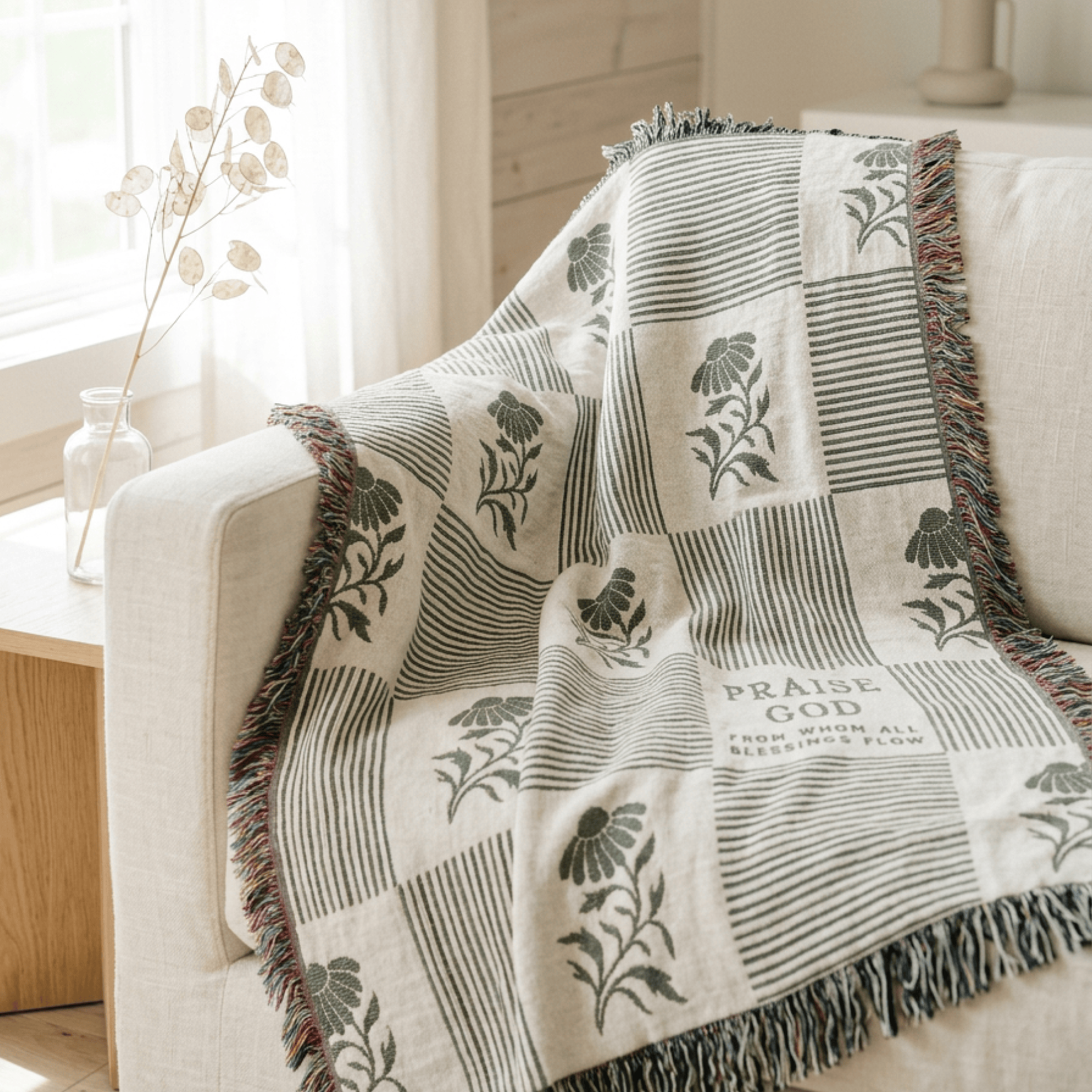 A cream and olive green throw blanket with a floral and striped patchwork pattern and fringed edges, draped over a light beige armchair in a bright, modern living room setting.