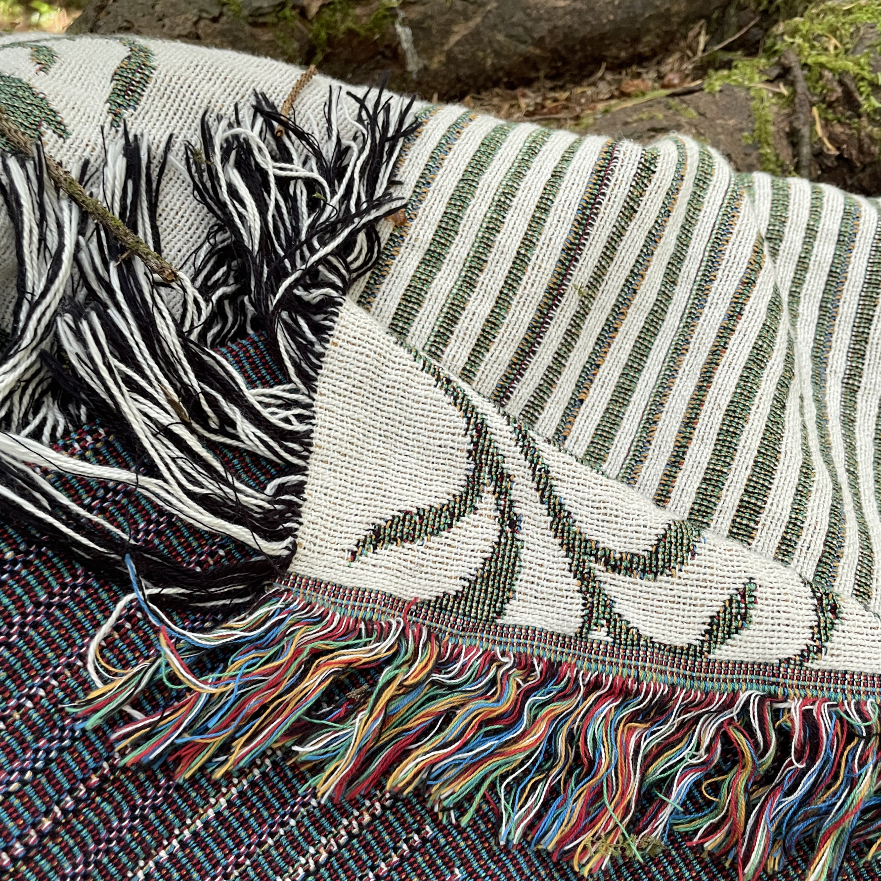 A close-up of a woven textile throw blanket with black and white fringe, striped green and white sections, and multicolored tassels, draped outdoors over another patterned blanket.