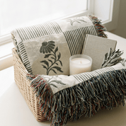 A woven basket on a windowsill holds a folded cream throw blanket with dark green floral and striped patterns, multicolor fringe, a white candle, and a small linen book or journal.