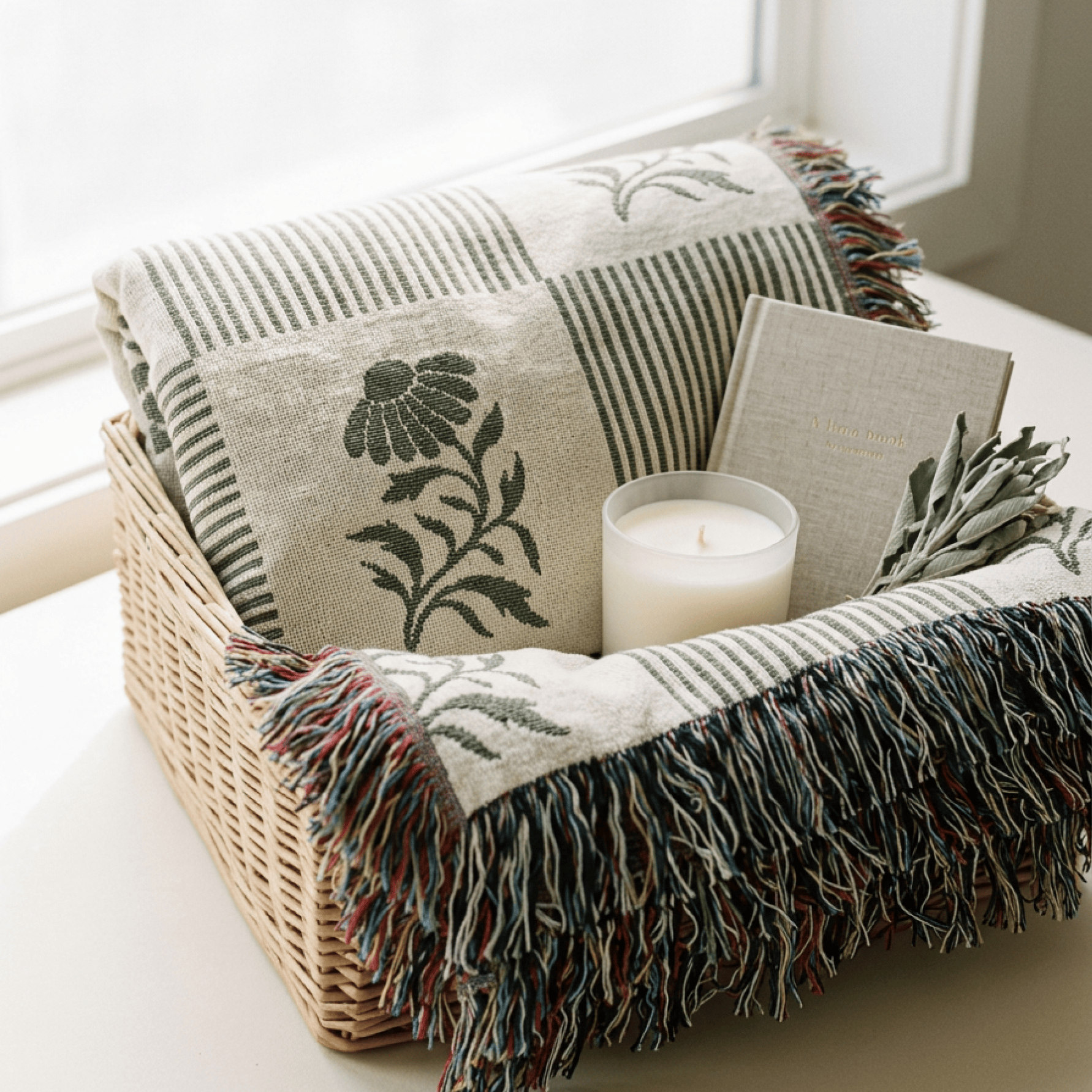 A woven basket on a windowsill holds a folded cream throw blanket with dark green floral and striped patterns, multicolor fringe, a white candle, and a small linen book or journal.