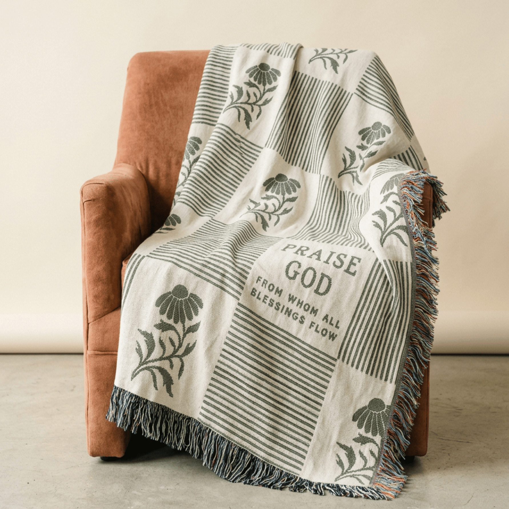 A cream and olive green patterned throw blanket with fringe, draped over a rust-brown armchair against a neutral beige wall.