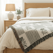 A woven throw blanket with a patchwork design of stripes, floral motifs, and fringe trim, styled on a light wood bed in a bright, modern bedroom.