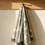 A patterned woven throw blanket with fringe is hanging from a metal hook on a wooden wall rod against a textured off-white wall. The blanket has a cream base with dark gray stripes, botanical motifs, and a Bible verse printed on it.