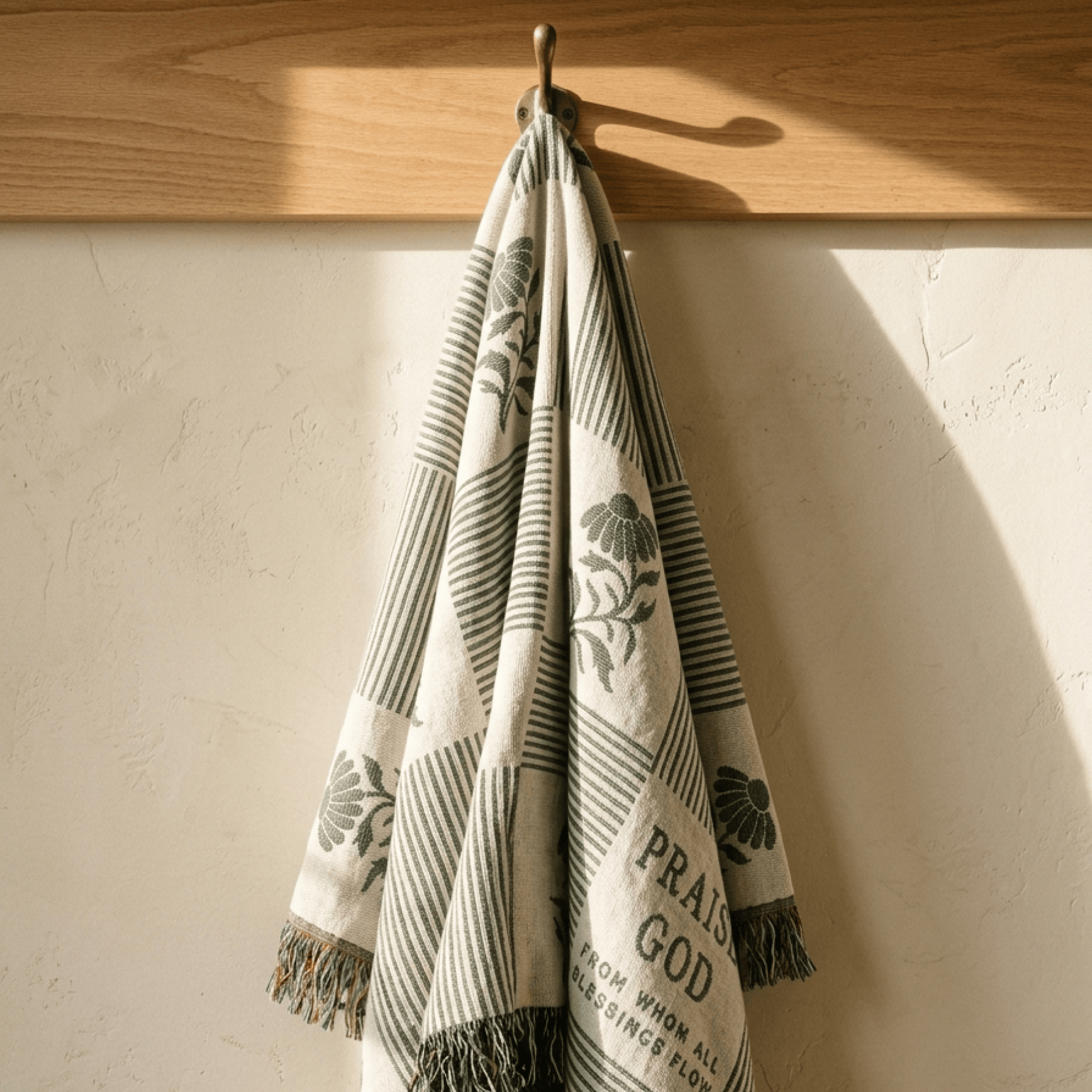 A patterned woven throw blanket with fringe is hanging from a metal hook on a wooden wall rod against a textured off-white wall. The blanket has a cream base with dark gray stripes, botanical motifs, and a Bible verse printed on it.