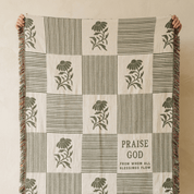 A checkered throw blanket with olive green floral and striped patterns on a cream background, held up by two hands against a neutral beige wall. The blanket features fringe on the sides and the text 'PRAISE GOD FROM WHOM ALL BLESSINGS FLOW' printed on one of the squares.