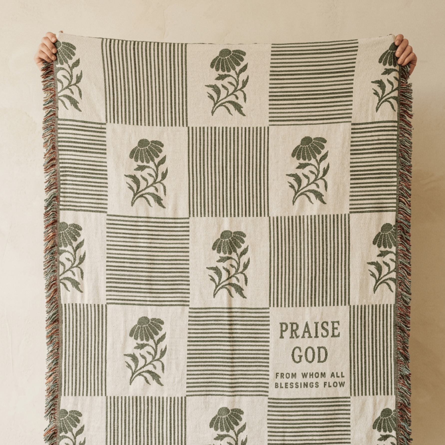 A checkered throw blanket with olive green floral and striped patterns on a cream background, held up by two hands against a neutral beige wall. The blanket features fringe on the sides and the text 'PRAISE GOD FROM WHOM ALL BLESSINGS FLOW' printed on one of the squares.