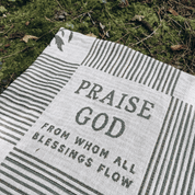 A woven outdoor blanket with the words 'PRAISE GOD FROM WHOM ALL BLESSINGS FLOW' laid on mossy ground.