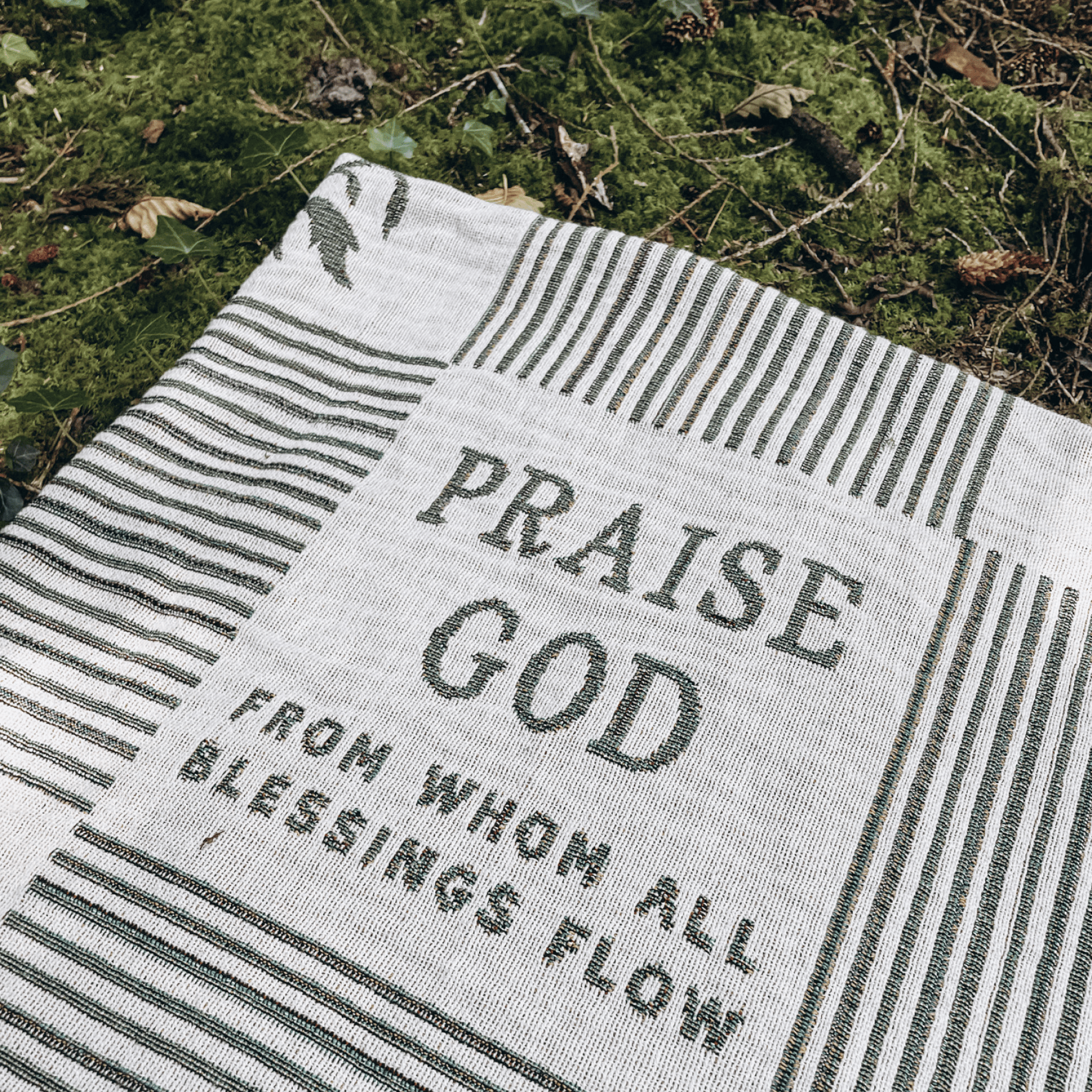 A woven outdoor blanket with the words 'PRAISE GOD FROM WHOM ALL BLESSINGS FLOW' laid on mossy ground.