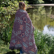 Person wrapped in a purple blanket with floral patterns and 'LORD' text, standing by a pond.