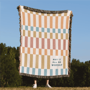 Checkered blanket with text 'May it all be worship' on a grassy field with trees in the background