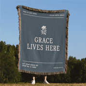 Blanket with 'Grace Lives Here' text and design, held up outdoors against a blue sky and trees.