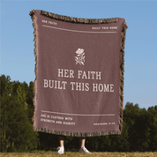 Brown blanket with text and flower design, held by a person in a grassy area with trees and blue sky.