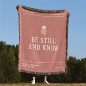Blanket with 'Be Still and Know' text and flower design in a natural setting