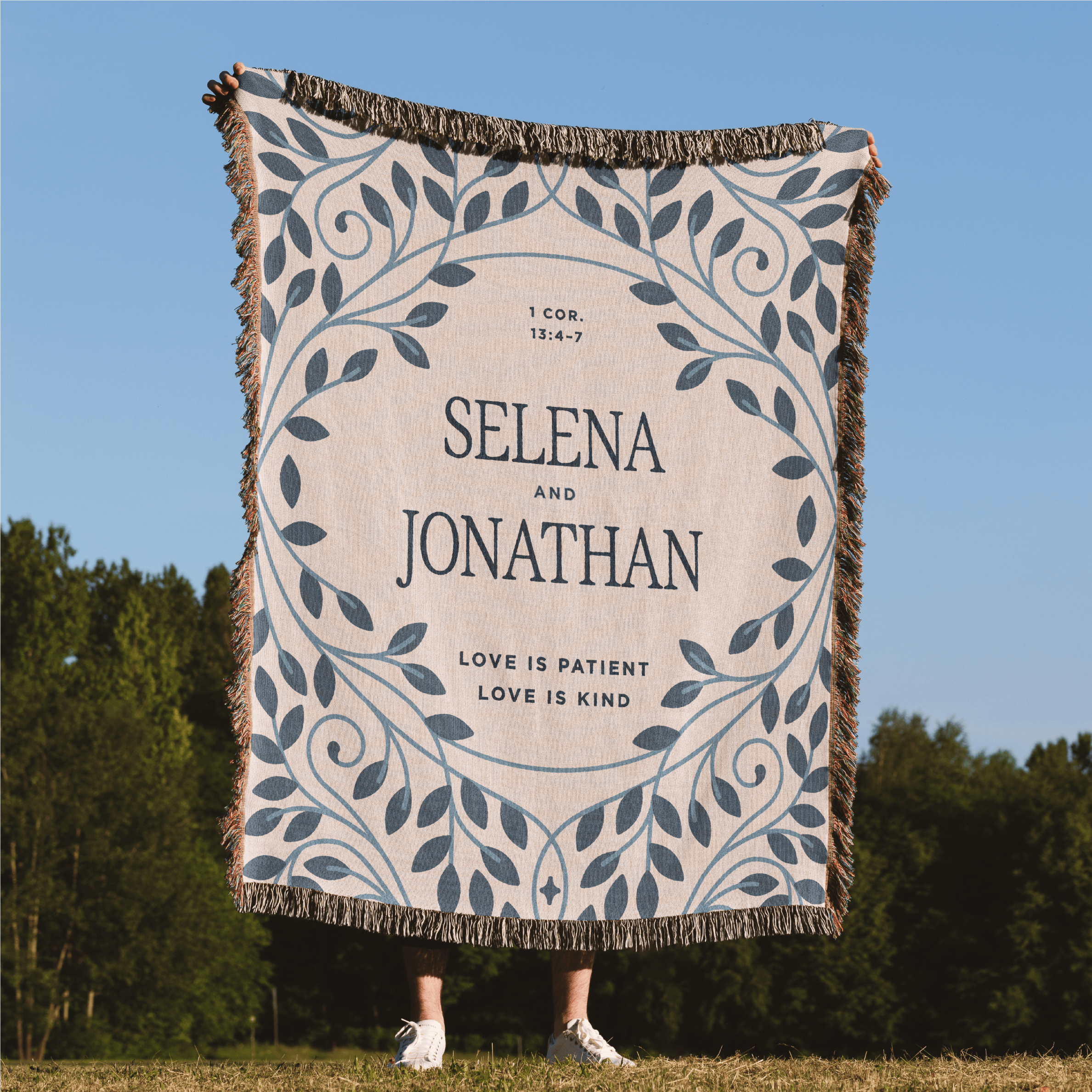 Personalized blanket with names and text, displayed outdoors against a blue sky and green trees.