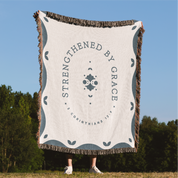 Blanket with 'Strengthened by Grace' text and design, held up outdoors.