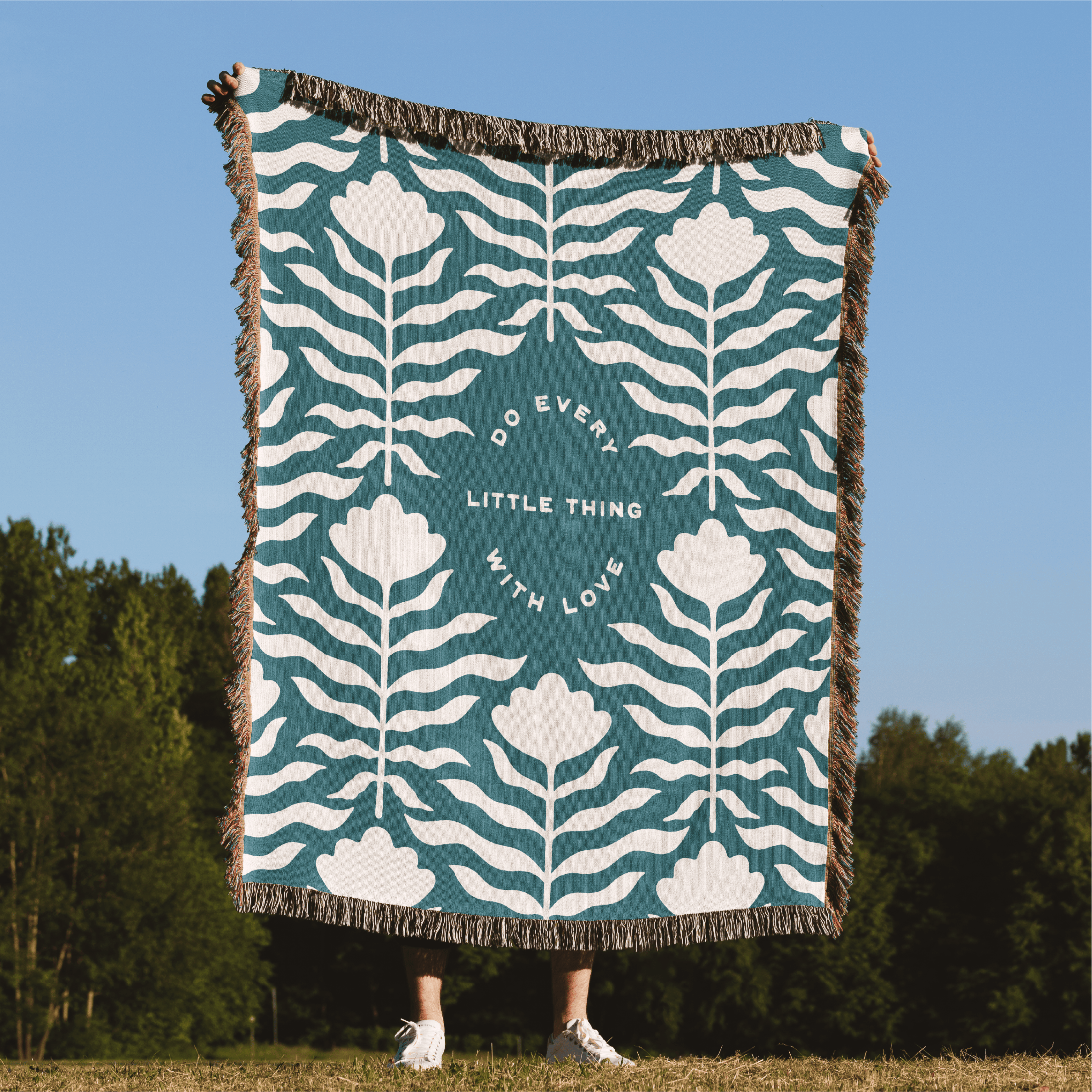 Blanket with floral pattern and text on a grassy field with trees in the background