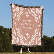 Blanket with leaf pattern and text 'Peace be to this house Luke 10:25' held by a person outdoors.