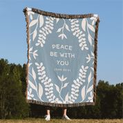 Blanket with 'Peace Be With You' text and leaf patterns in a natural setting