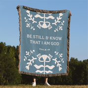 Blue blanket with white text and designs, featuring 'Be Still & Know That I Am God' from Psalm 46, against a natural background.