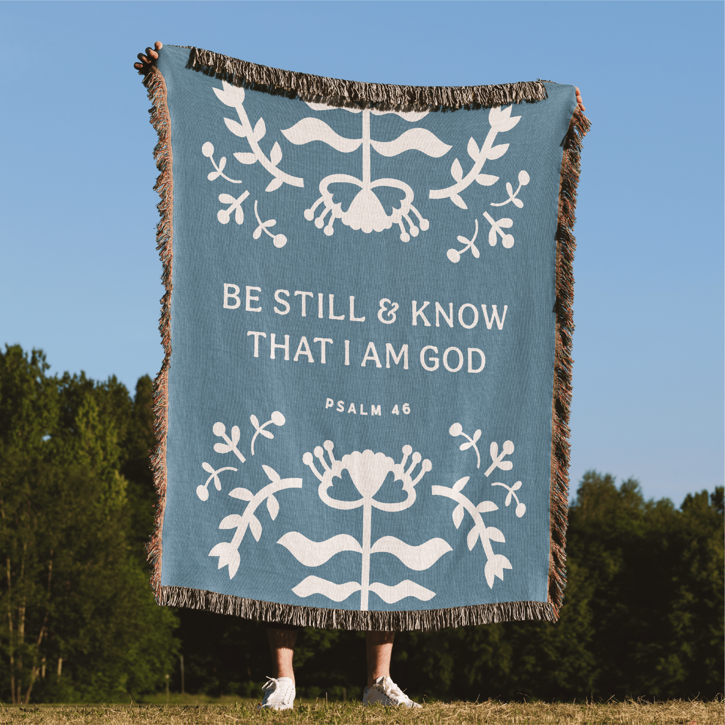 Blue blanket with white text and designs, featuring 'Be Still & Know That I Am God' from Psalm 46, against a natural background.