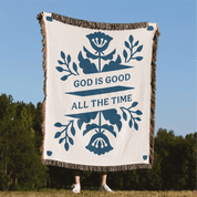 Blanket with floral design and 'God is Good All the Time' text against a blue sky and green trees background