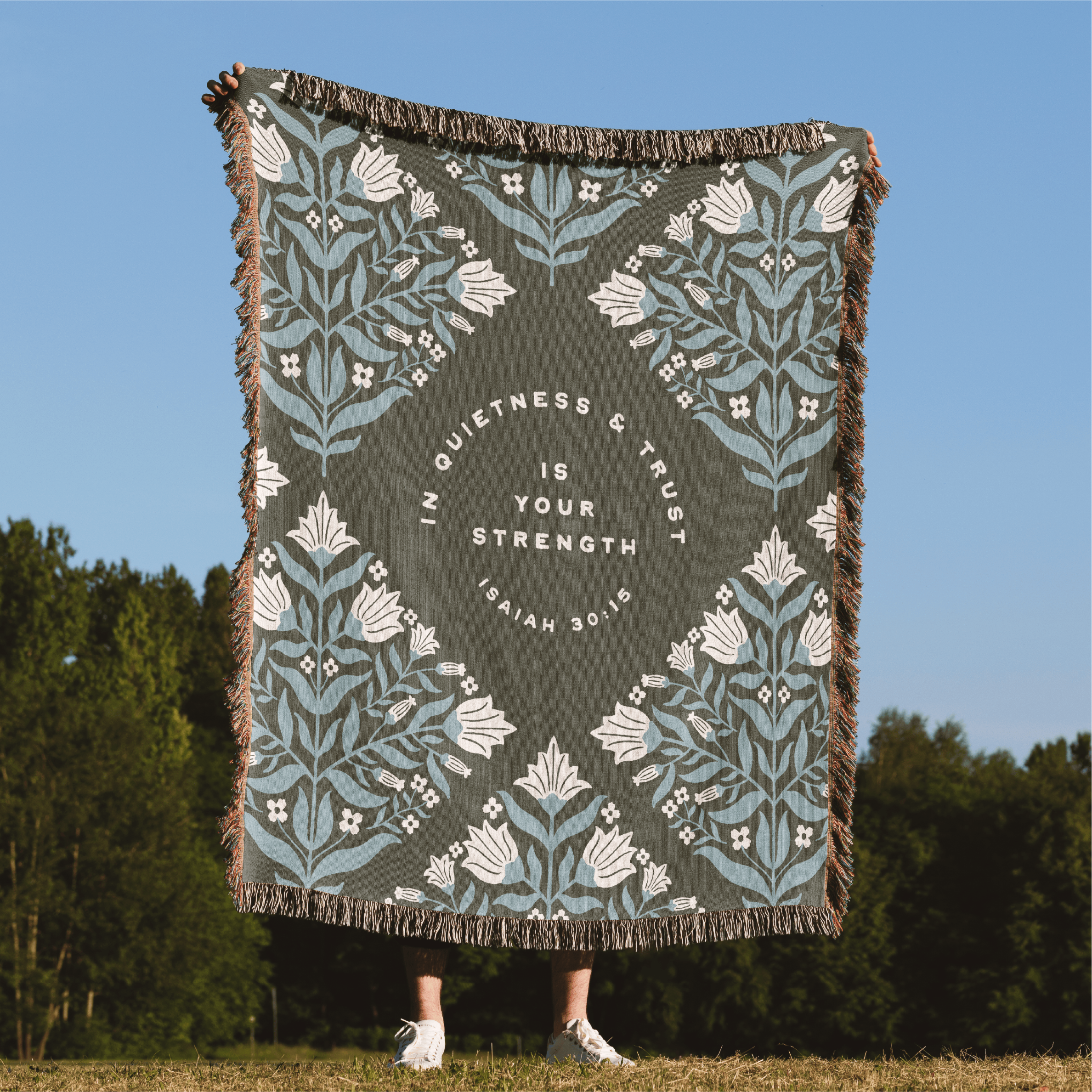 Decorative blanket with floral patterns and text on a grassy field with trees in the background