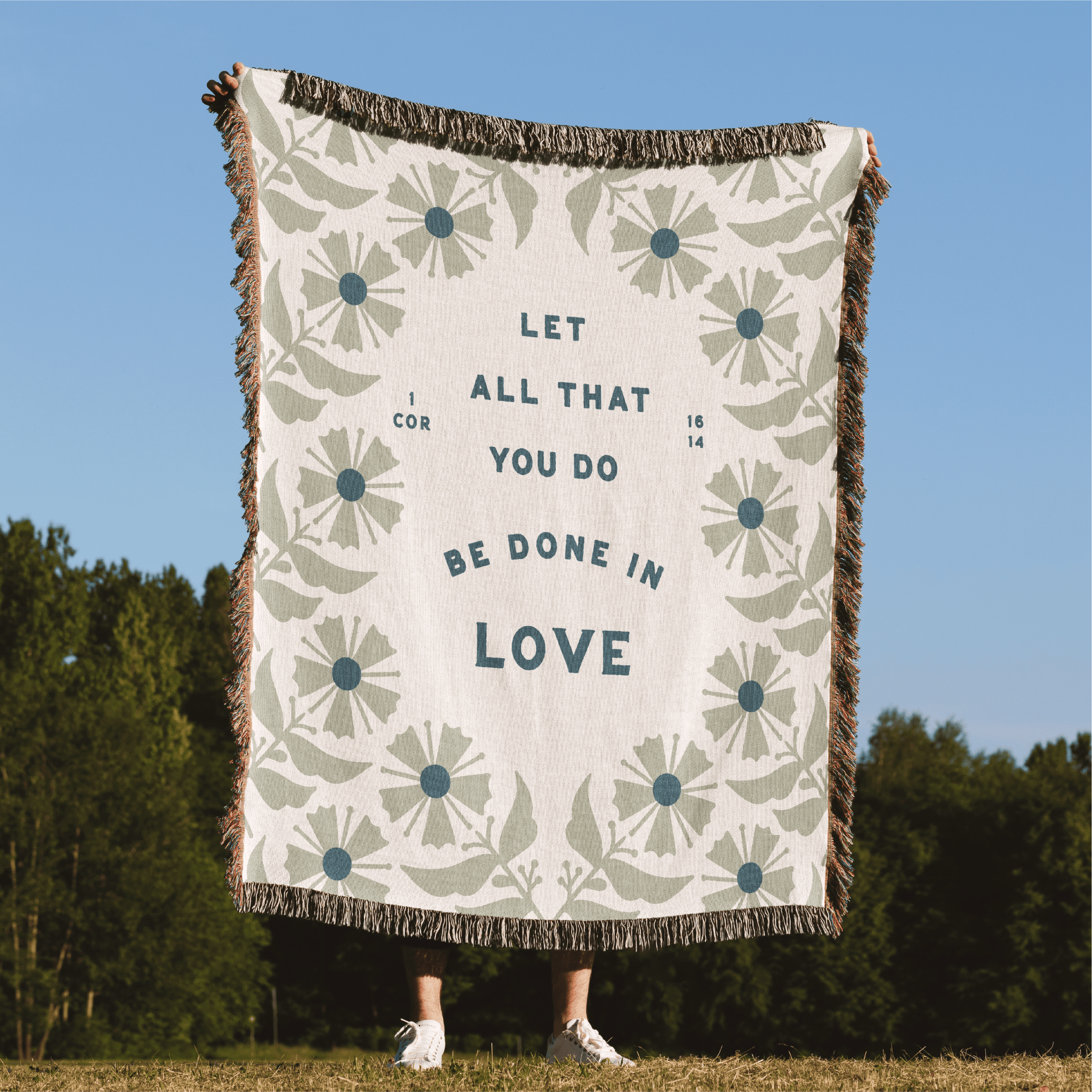 Blanket with floral pattern and text 'Let all that you do be done in love' against a blue sky and green trees background