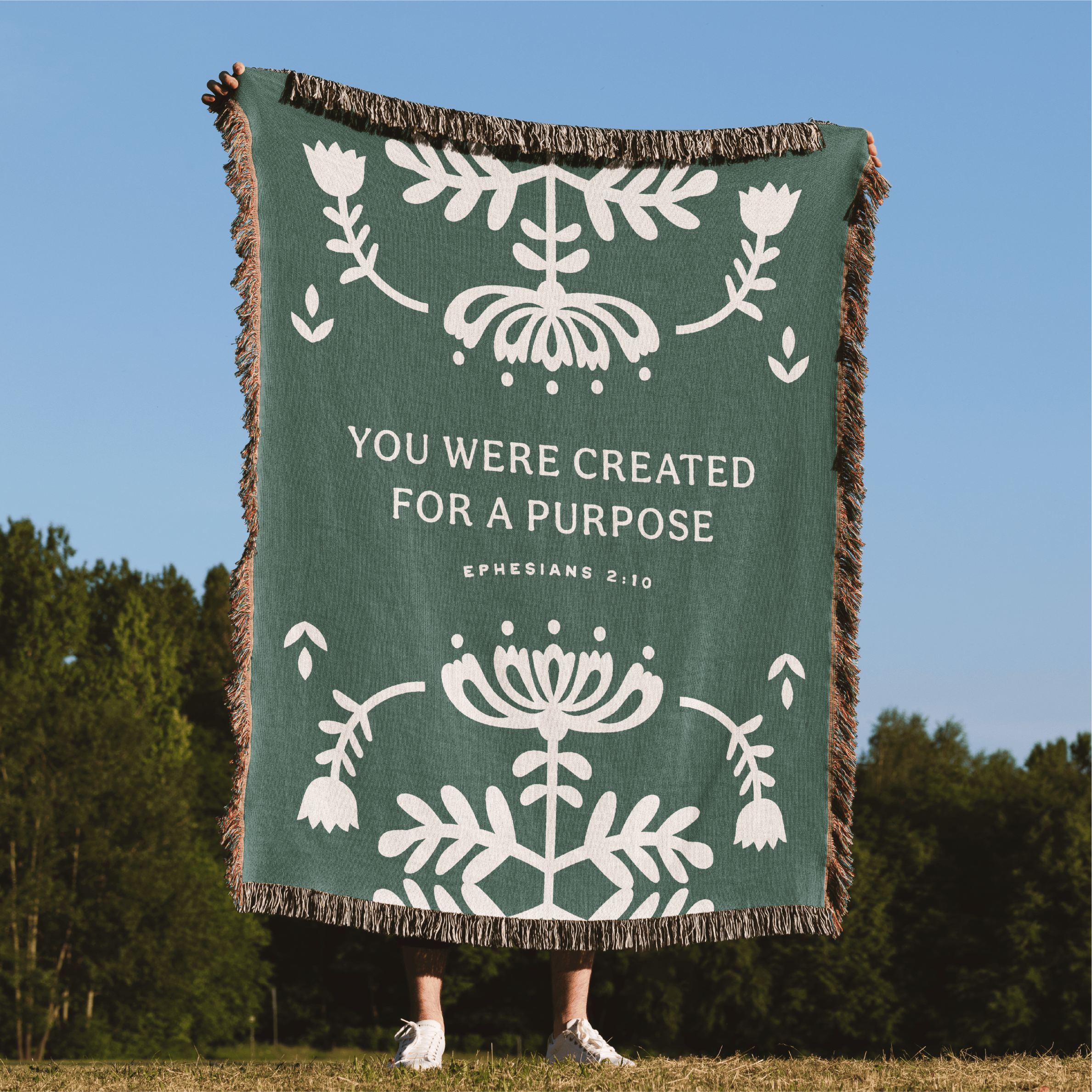 Green blanket with white text and floral patterns on a grassy field with trees and blue sky.