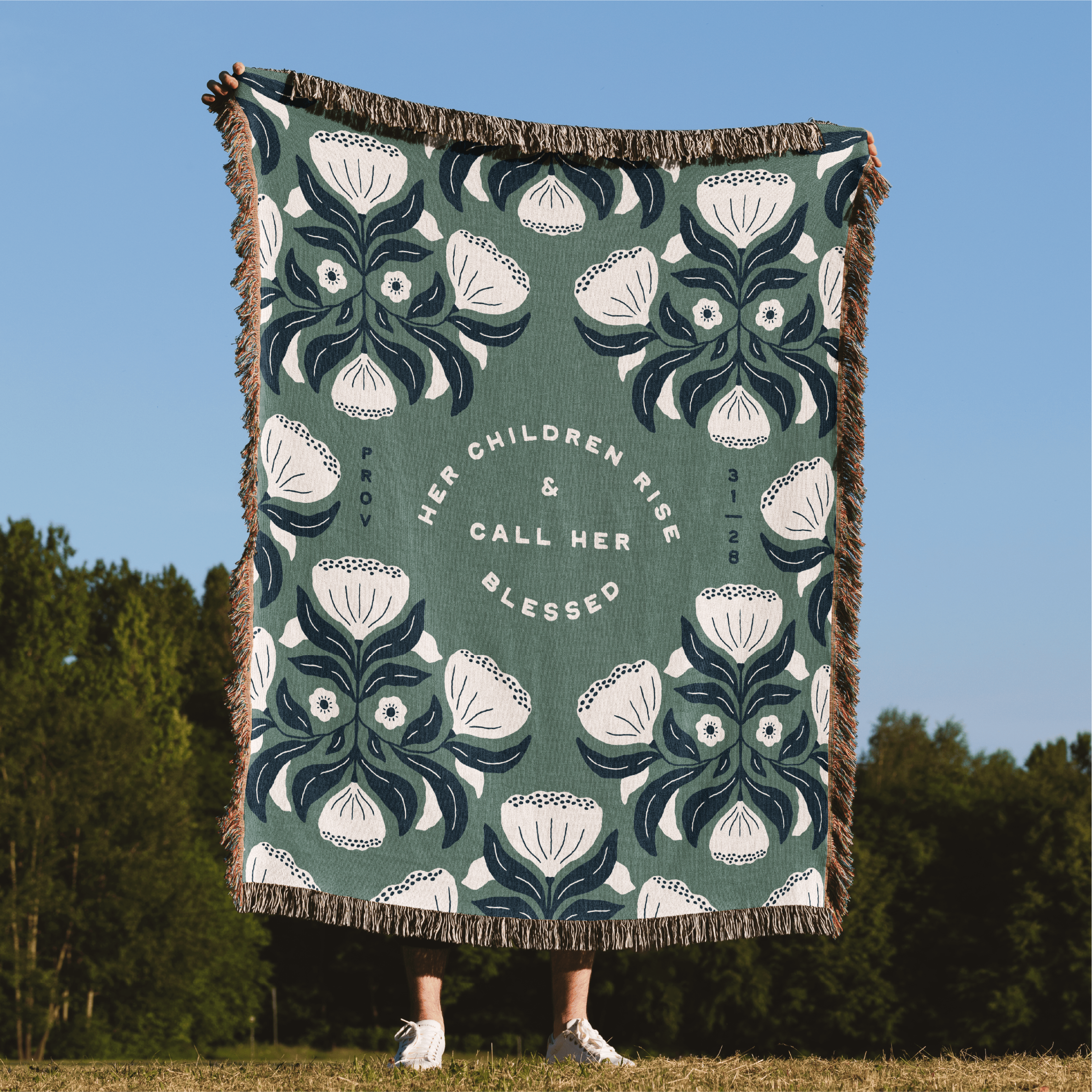 Decorative blanket with floral pattern and text, displayed outdoors against a blue sky and trees.