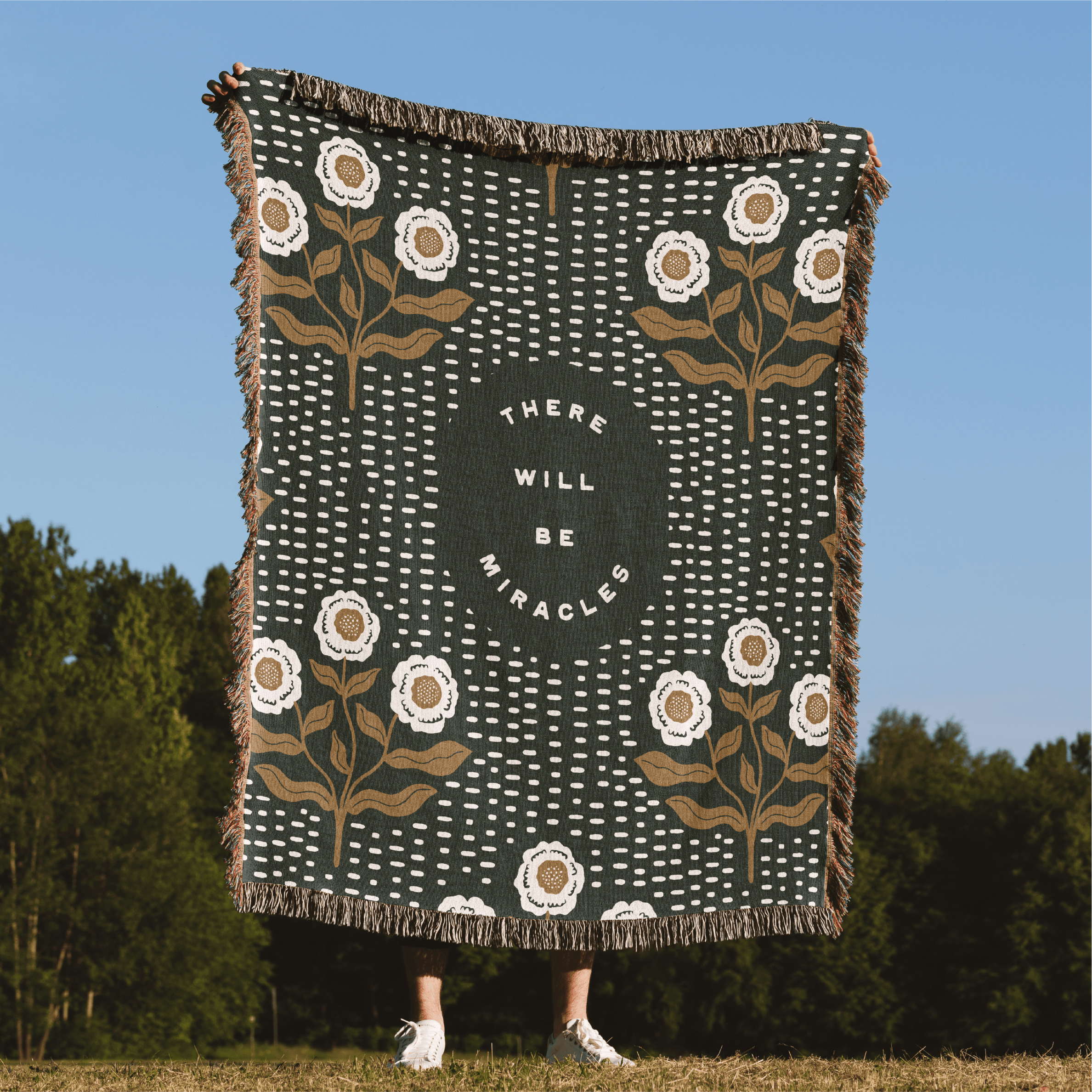 Decorative blanket with floral patterns and 'There will be miracles' text, held by a person outdoors.