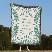 Blanket with green leaf pattern and text 'His mercies are new every morning' on a grassy field with trees and blue sky.
