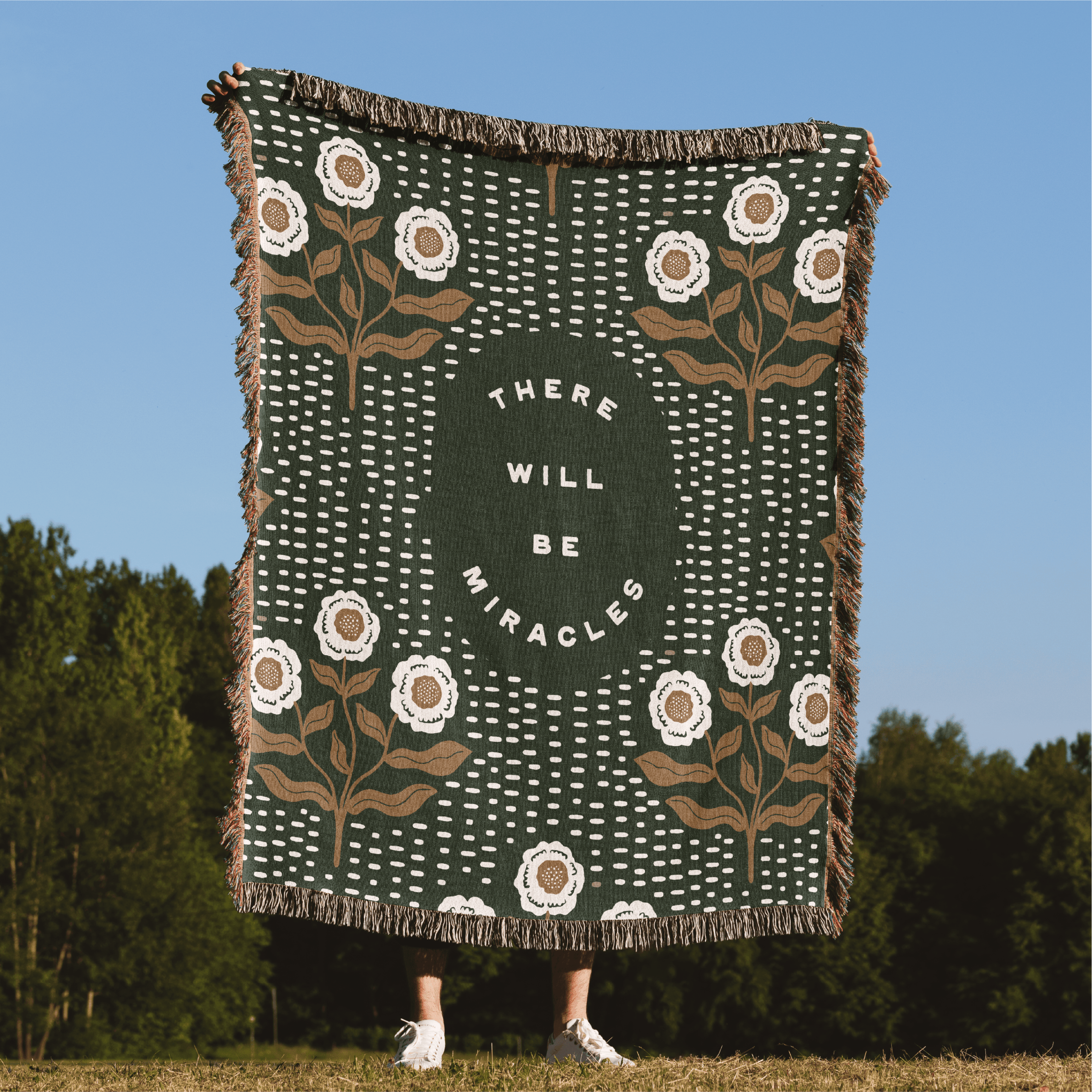 Decorative blanket with floral patterns and 'There will be miracles' text, displayed outdoors.