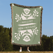 Green blanket with white floral design and text 'I was lost now I'm found' against a blue sky and green trees background.