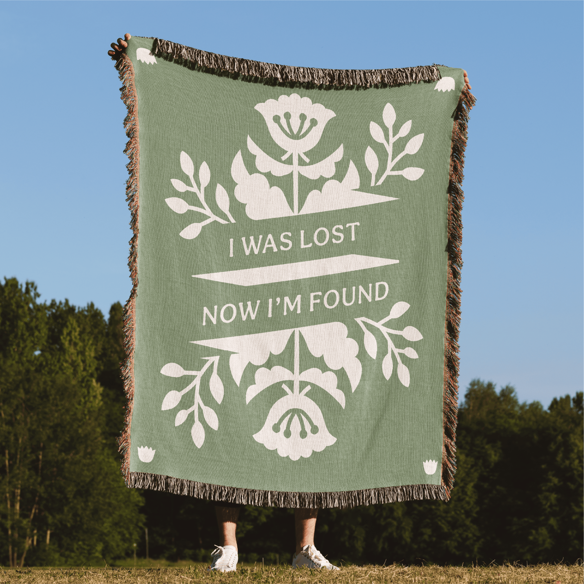 Green blanket with white floral design and text 'I was lost now I'm found' against a blue sky and green trees background.