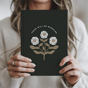 Person holding a black notebook with floral design and 'There will be miracles' text.