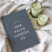 Gray book with 'Her Faith Carries Us' text on a beige blanket with two candles.